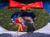 Two people in festive clothing sit inside a large holiday wreath adorned with lights and a big red bow.
