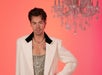 A lifelike wax figure of Harry Styles dressed in a stylish white blazer over a sparkling gold top, accessorized with a pearl necklace and a cross pendant, set against a pink background with a chandelier above.