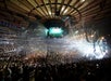 A large crowd fills an indoor arena with bright stage lights and a screen displaying visuals. The wooden ceiling and multilevel seating are visible.