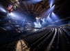 An empty arena with lit stage and rows of seating, illuminated by bright overhead spotlights. An American flag hangs in the upper left.