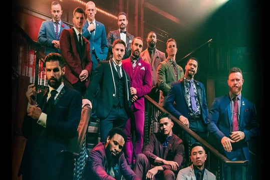 A group of men dressed in colorful suits pose on a staircase under dramatic lighting in a formal setting.