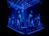 A group of male dancers wearing jeans and sneakers perform on a stage under blue lighting, surrounded by illuminated stage fixtures.