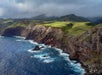 A rugged coastline with steep cliffs and rocky shores meets a choppy blue ocean under a cloudy sky. Green grass and hills stretch across the landscape.