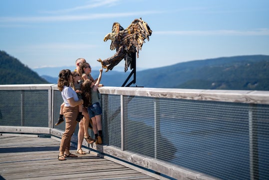 A family stands on a boardwalk overlooking a scenic landscape with mountains. They examine an eagle sculpture perched on the railing.