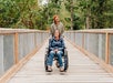 Two people on a wooden bridge; one is in a wheelchair smiling, the other stands behind, also smiling. Trees surround the path.