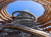 View from below of a spiral slide and metal staircase inside an open-roof, circular wooden structure against a clear blue sky.