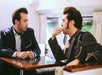 Two men with similar hairstyles and outfits sit across from each other at a table in a room with a window and curtains, one looking at the other who is resting his hand on his chin.