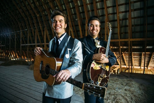 Two men in vintage-style suits stand in a wooden barn, each holding a guitar and smiling at the camera.