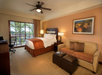 Hotel room with a double bed, sofa, two ottomans, desk with chair, ceiling fan, and a lamp. A framed picture hangs on the wall, and a window overlooks palm trees.