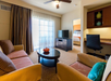 Hotel suite living area with two sofas, a round coffee table, a TV, and a desk chair. A doorway leads to a bedroom. Large window with curtains on the left.