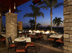 Outdoor restaurant patio at sunset with tables set for dining, surrounded by stone pillars and lit fire features. Palm trees and a clear sky in the background.