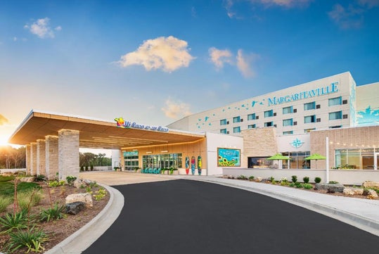 Exterior view of a Margaritaville resort hotel with a covered entrance, colorful signage, and landscaping under a partly cloudy sky.