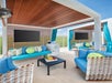Outdoor cabanas with wicker furniture, blue cushions, and decorative pillows are arranged around small tables with fruit platters under wooden roofs, with bright blue curtains and grass in the background.