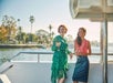 Two women standing on a boat deck, holding drinks and smiling, with water, palm trees, and buildings visible in the background on a sunny day.