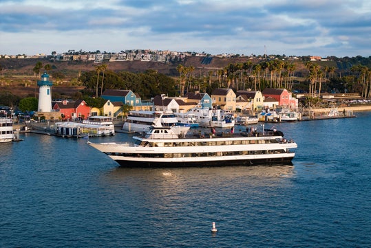 A large white and black yacht sails near a marina with colorful buildings, a lighthouse, and palm trees along the shoreline under a partly cloudy sky.