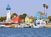 Colorful houses, a blue and white lighthouse, and several boats line the waterfront under a clear sky.
