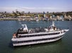 A large white yacht with people on board sails near a marina with docks, palm trees, and buildings in the background on a sunny day.