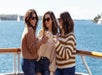 Three women wearing sweaters and sunglasses stand on a boat deck, holding drinks and smiling, with water and sailboats visible in the background.