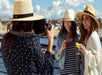 Two women in hats holding drink posing for another woman in a hat to take their photo on bright sunny day.
