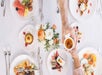 A white table full of plates of colorful food with hands passing a small dish in a ramekin across the table.