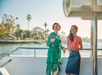 Two women stand on a yacht deck with drinks, overlooking a calm waterway lined with palm trees.