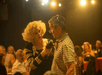 Performer with curly hair holds a microphone to a man in a striped shirt in a dimly lit room with audience in the background.