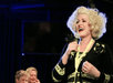 Person with curly blond hair holding a microphone, wearing a black and yellow outfit, smiles while speaking on stage. Two people visible in the background, partially out of focus.