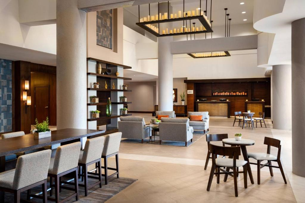 Modern hotel lobby with seating areas, a long table, and reception desk. Neutral colors and minimalist decor.