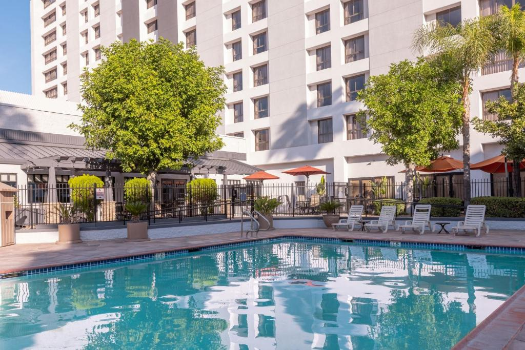 Outdoor hotel pool surrounded by lounge chairs and trees, with a multi-story building in the background.