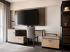 Hotel room desk area with a wall-mounted TV, a chair, and a lamp. A bench and storage cabinet are also visible. Neutral tones and modern design.