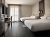 Modern hotel room with two double beds, a desk with a chair, a TV, and a large window with sheer curtains.