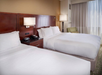 Hotel room with two neatly made double beds, a wooden headboard, a nightstand with a phone and lamp, and a window with sheer curtains.