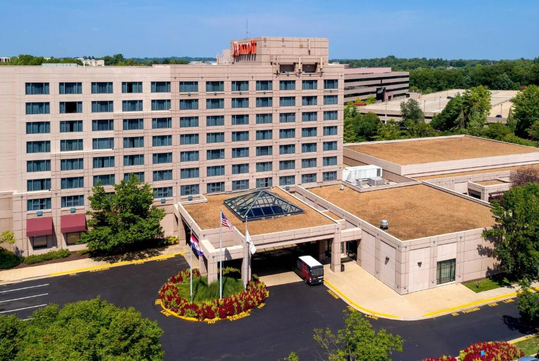 Aerial view of a multistory hotel with a landscaped entrance and parking area, surrounded by trees and other buildings in the background.