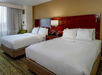 Hotel room with two neatly made double beds, a wooden headboard, a window with sheer curtains, and a green chair in the corner.