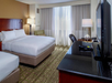 Hotel room with two double beds, a TV on a black dresser, a lamp on a desk, and natural light coming through curtains.