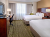 Hotel room with two double beds, a desk with a chair, a television on a cabinet, and a window with sheer curtains. The carpet is patterned, and the walls are light-colored.
