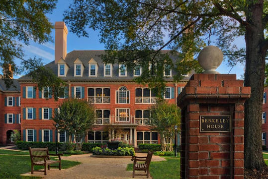 Red brick building with white trim and rows of windows, surrounded by trees and benches. A brick pillar in the foreground has a plaque reading "Berkeley House.