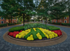 Circular garden with neatly trimmed hedges and colorful flower beds, surrounded by brick paths and red brick buildings, under a canopy of trees.