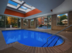 Indoor swimming pool with clear blue water, surrounded by windows revealing an outdoor view. The ceiling has glass panels. Steps lead into the pool on the right.