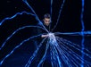 A man in dark clothing interacts with multiple glowing blue light beams projecting outward from his hands against a dark background.