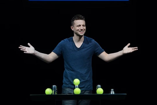 A man stands behind a table with three tennis balls and three metal cups, arms outstretched, smiling against a black background.