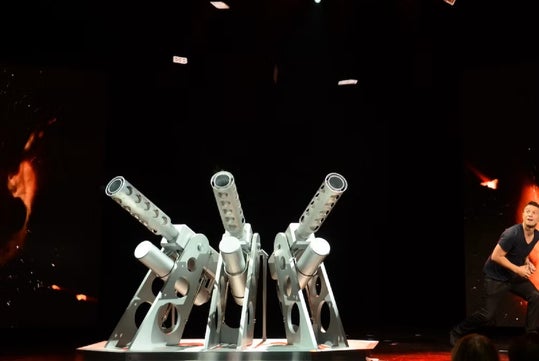 A man stands beside a large metallic structure with four angled cylindrical tubes, set on a stage with a dark background and fiery visuals.