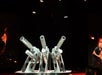 A man stands beside a large metallic structure with four angled cylindrical tubes, set on a stage with a dark background and fiery visuals.