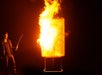 A person holding a sword stands next to a large rectangular object engulfed in flames against a dark background.