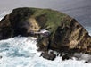 A helicopter flies near a rocky, grass-covered coastal cliff with waves crashing against the shore below.