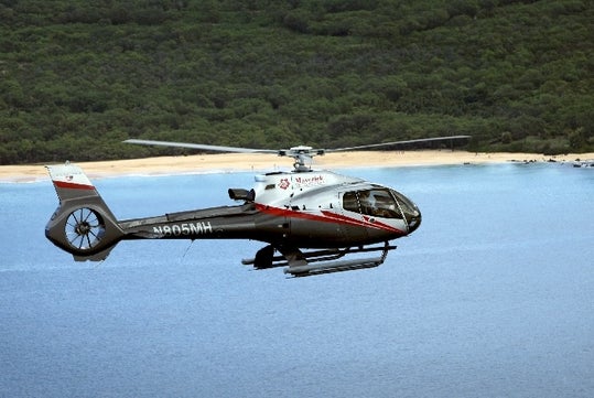 A silver and black Maverick airbus flying over the beautiful seas of Maui with the dense tree covered island in the background.