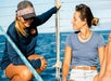 Two people on a boat, smiling and conversing. One wears a cap and sunglasses, sitting on the railing. The other wears a blue shirt and shorts, seated. Ocean background visible.