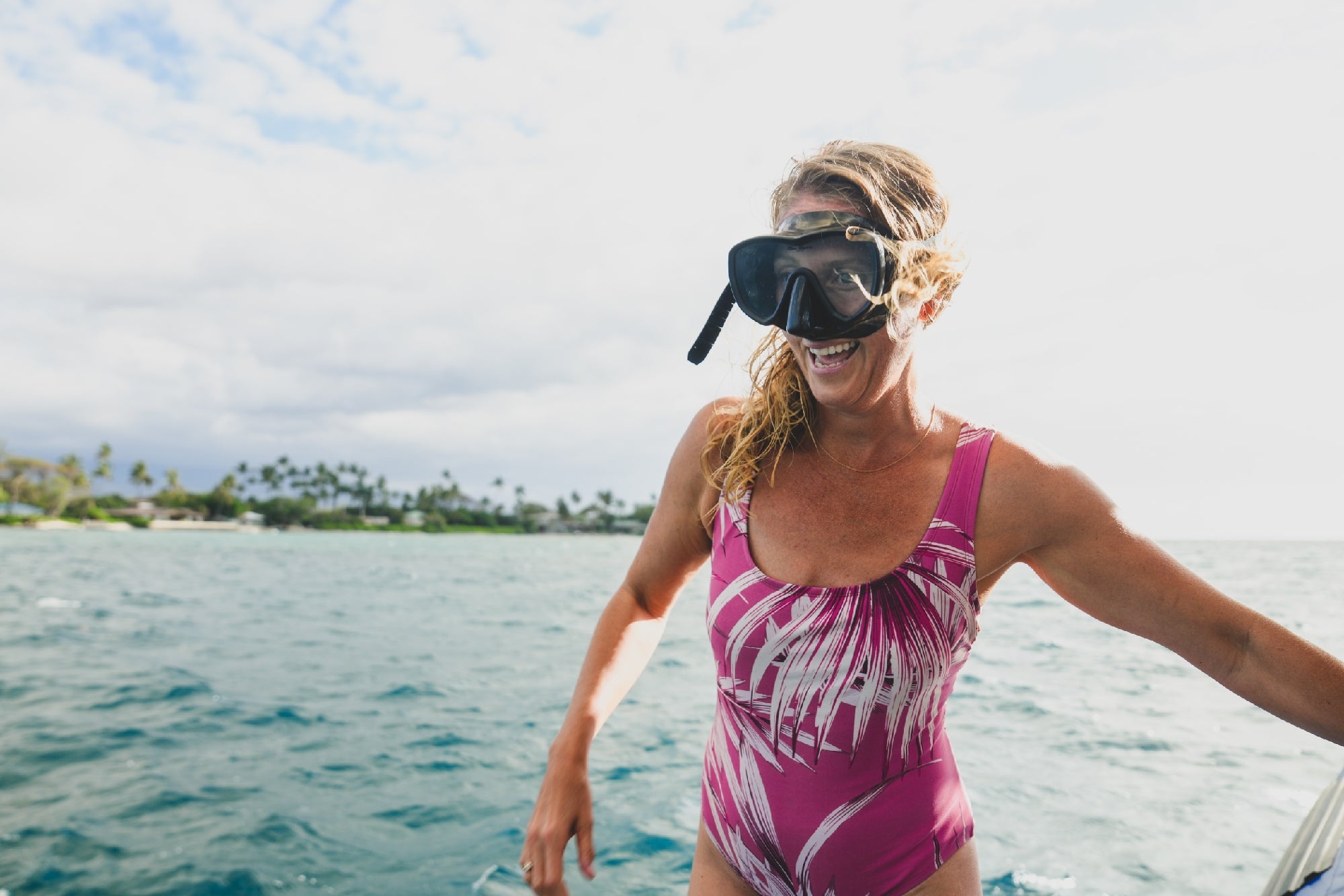 A woman in a pink patterned swimsuit and snorkel mask stands smiling by the ocean with water and a shoreline in the background.