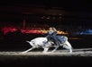 A woman in a dark, elaborate dress rides a white horse performing a bow during an indoor show with an audience in the background.