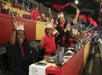 A group of people wearing crowns and red shirts enthusiastically wave flags and cheer inside an indoor arena with colorful seating.